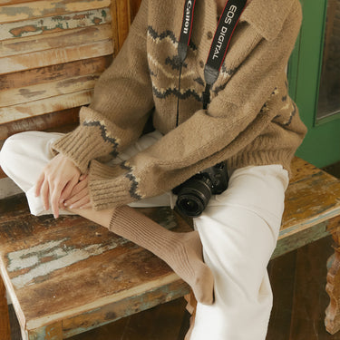 Person wearing a brown patterned sweater and white pants sitting on a wooden bench with a camera.